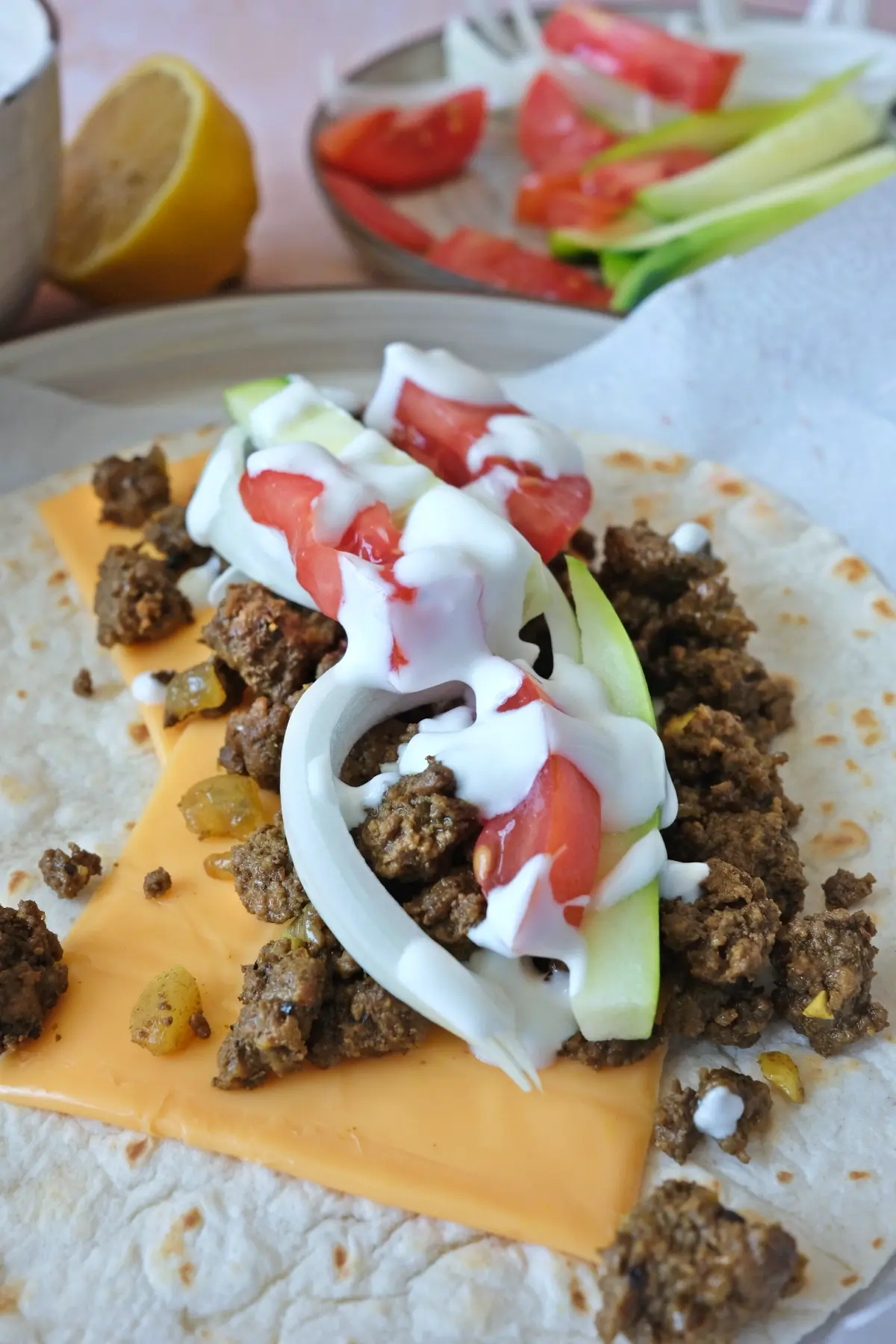 Open flour tortilla with melted American cheese, ground beef shawarma, cucumber sticks, tomato slices, and white onion strips layered before wrapping
