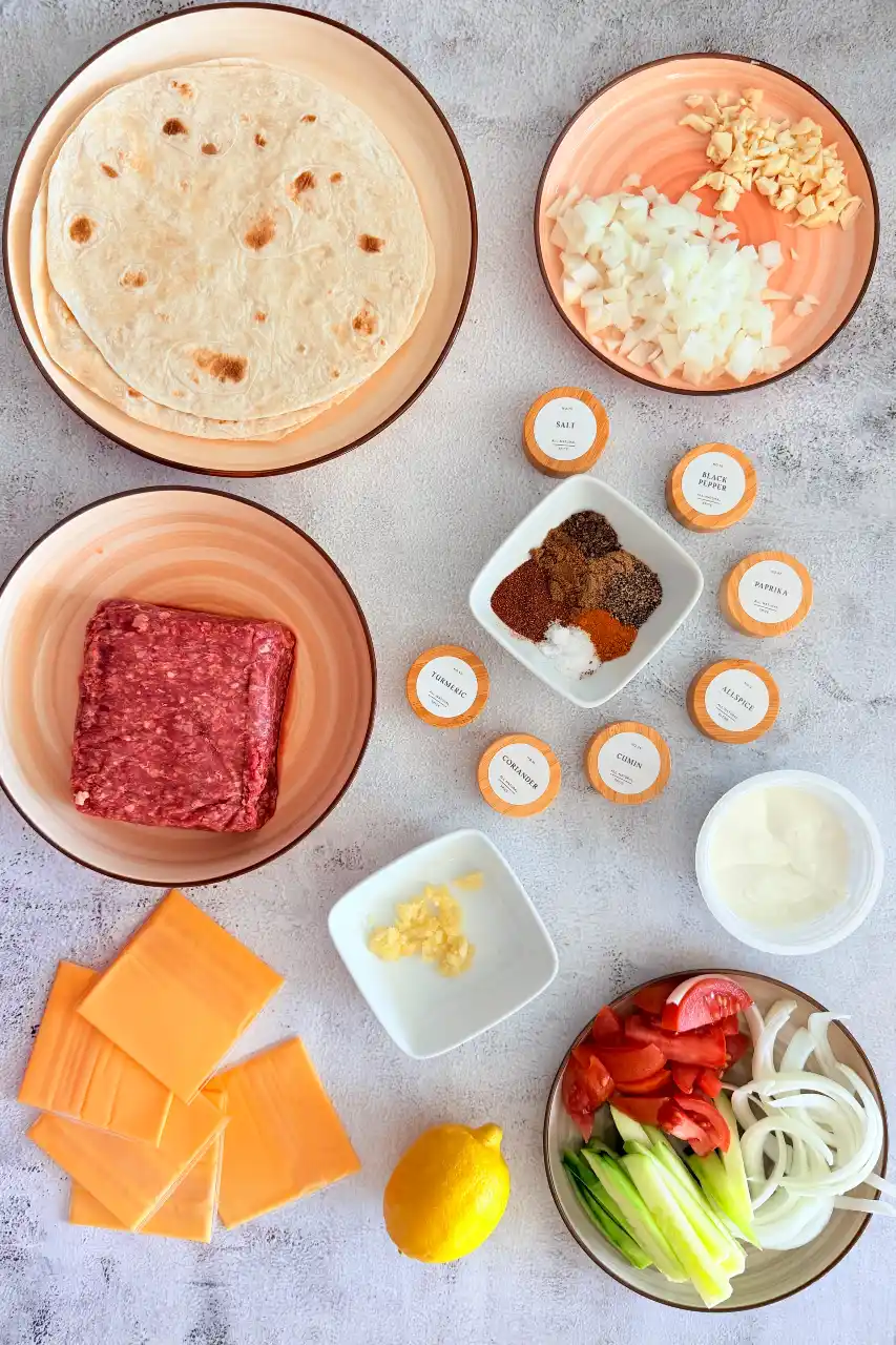 Overhead view of ground beef shawarma ingredients including flour tortillas, raw ground beef, sliced cheddar cheese, chopped onions, minced garlic, assorted spices in small bowls, yogurt, lemon, and fresh cucumber, tomato, and onion slices.