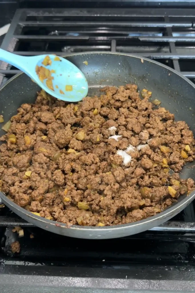 Cooked ground beef mixture in a skillet with a spoon sprinkling sugar over the top for final seasoning.