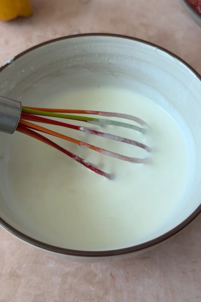 Creamy yogurt garlic sauce whisked until smooth in a bowl, with a whisk resting inside.