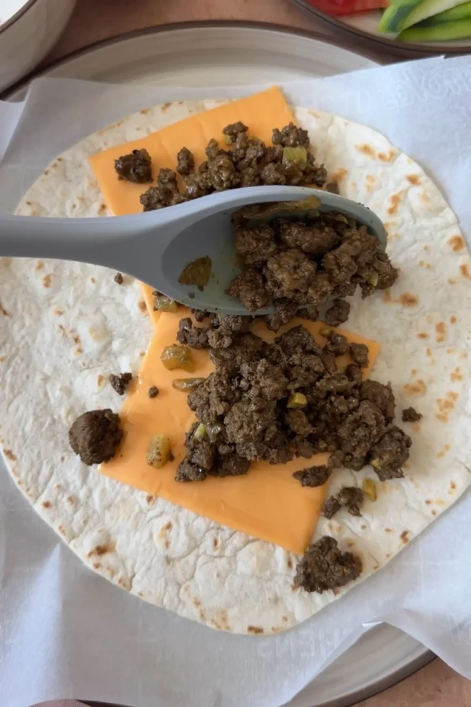 Seasoned ground beef spooned over sliced cheddar cheese on a tortilla, beginning the wrap assembly.