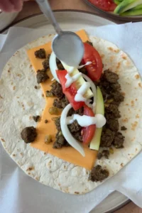 Creamy yogurt sauce drizzled over the beef, vegetables, and cheese on a tortilla before folding into a wrap.