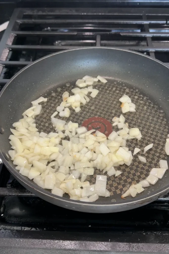 Diced white onions beginning to sauté in oil in a nonstick skillet on a stovetop, just starting to soften with slight translucency.
