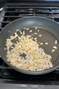 Chopped garlic added to sautéing onions in a pan, with pieces lightly turning golden as they cook together in oil.