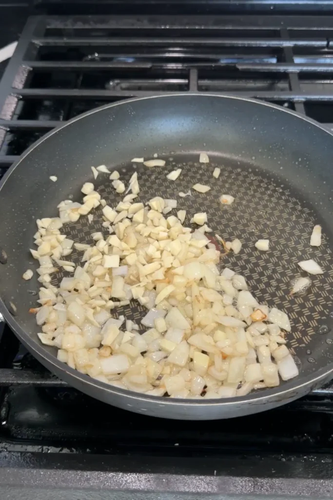 Chopped garlic added to sautéing onions in a pan, with pieces lightly turning golden as they cook together in oil.