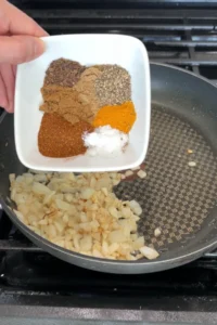 A small bowl of mixed spices including paprika, cumin, coriander, turmeric, black pepper, and salt being tipped into sautéed onions and garlic in a skillet.