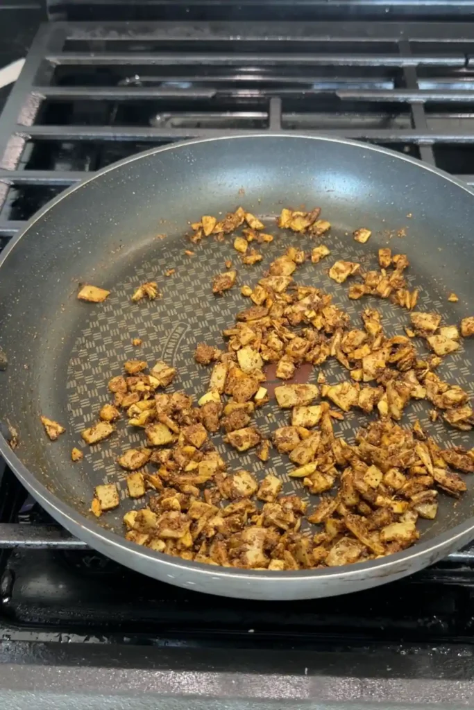 Onions and garlic coated in toasted spices in a skillet, darkened and fragrant with visible seasoning clinging to the pieces.