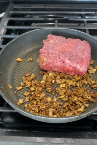 Block of raw ground beef placed into a skillet over spiced onions and garlic, beginning the cooking process.