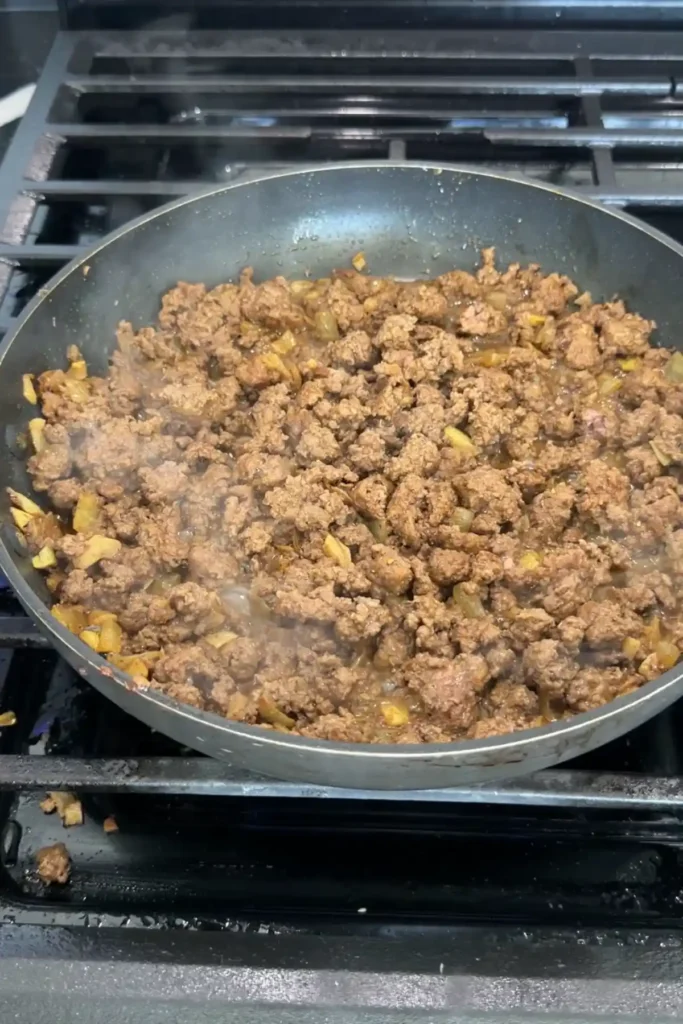 Ground beef fully browned and crumbled in a skillet with onions and spices, releasing steam as it cooks.