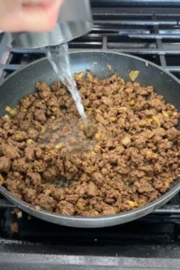 Water being poured into browned ground beef in a skillet to loosen the mixture and create a slightly saucy texture.