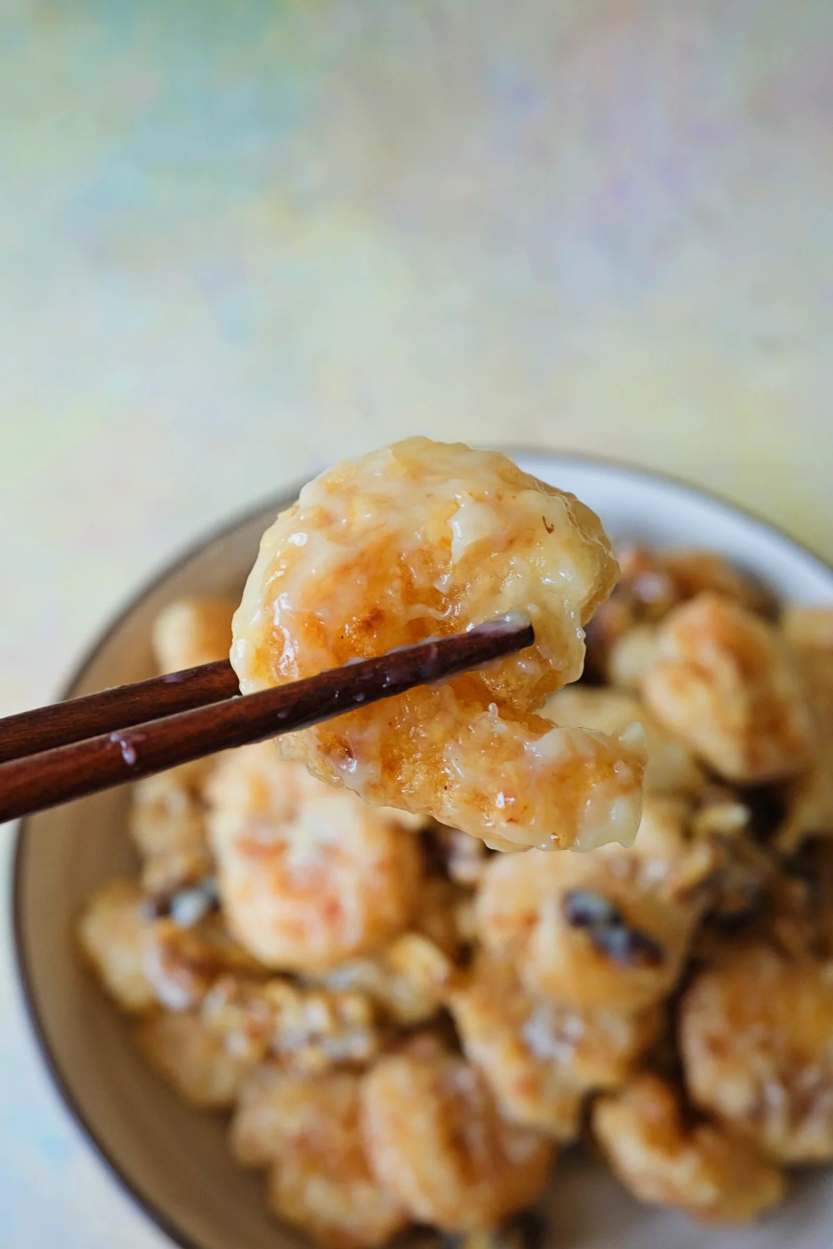 Chopsticks holding one piece of honey walnut shrimp above a blurred bowl, showing the thick creamy coating and crispy fried texture underneath.