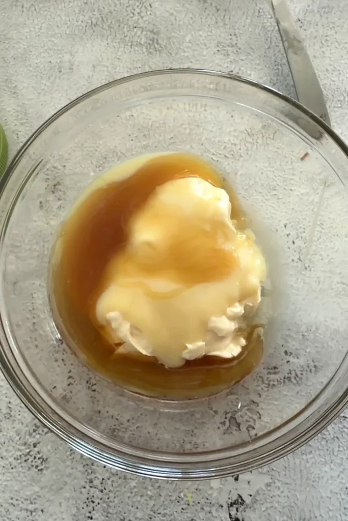 Mayonnaise and honey in a glass bowl before mixing, placed on a light textured countertop with a knife nearby