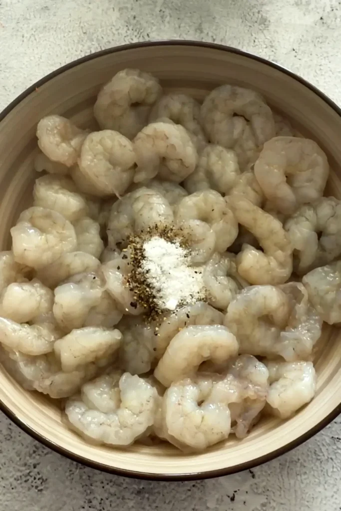 Raw peeled shrimp in a bowl seasoned with salt, pepper, and white powder on top before mixing