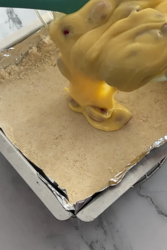 Lemon raspberry filling being poured over the shortbread crust in a lined baking pan.