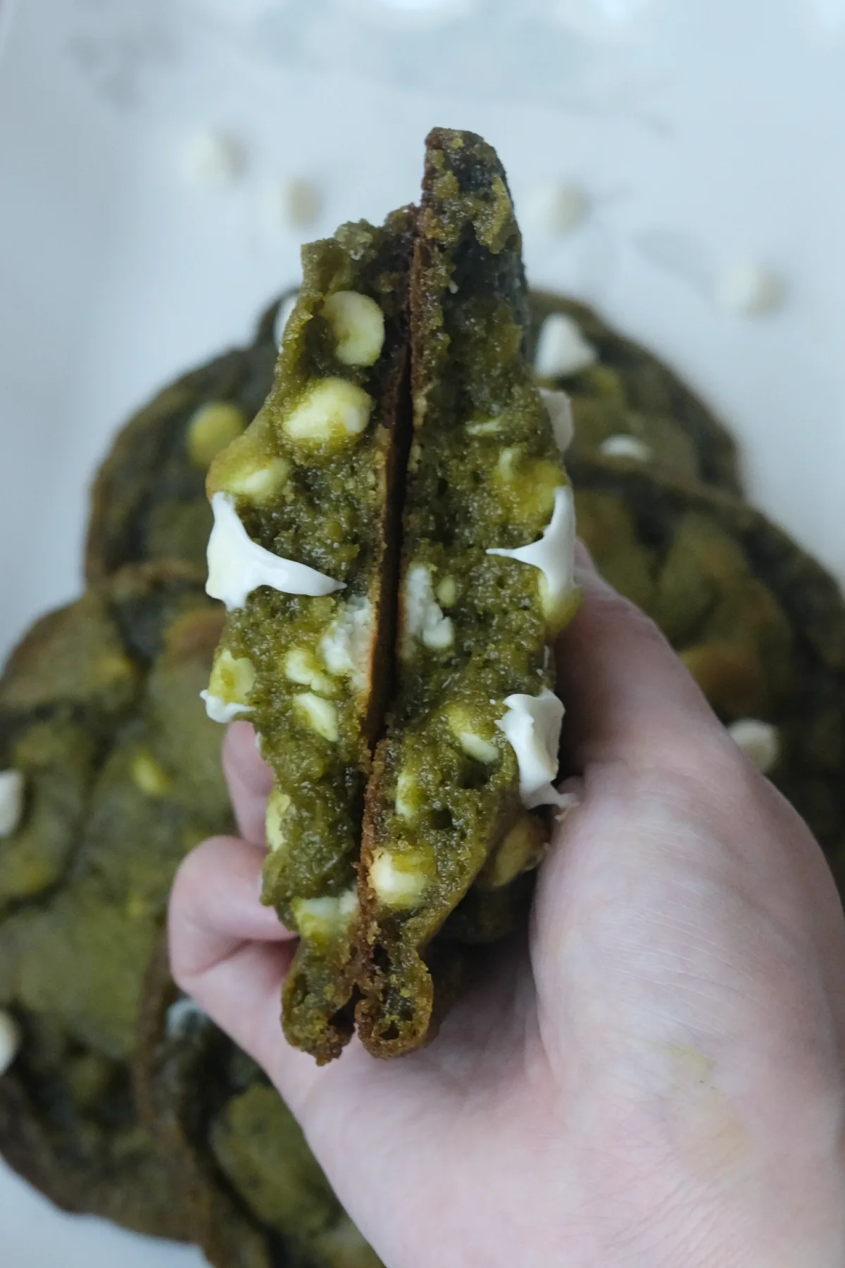 Hand holding a matcha white chocolate cookie split in half to show the soft, chewy center and white chocolate pieces.