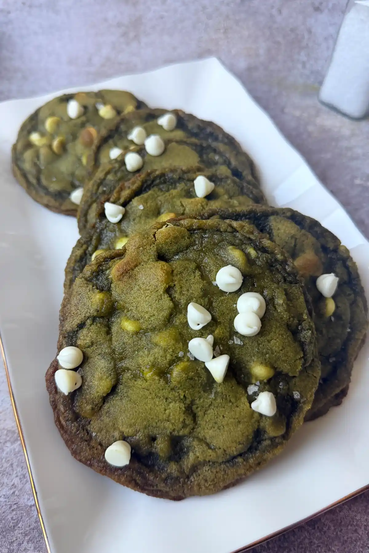 Six matcha white chocolate cookies arranged on a white rectangular plate with extra white chocolate chips on top.