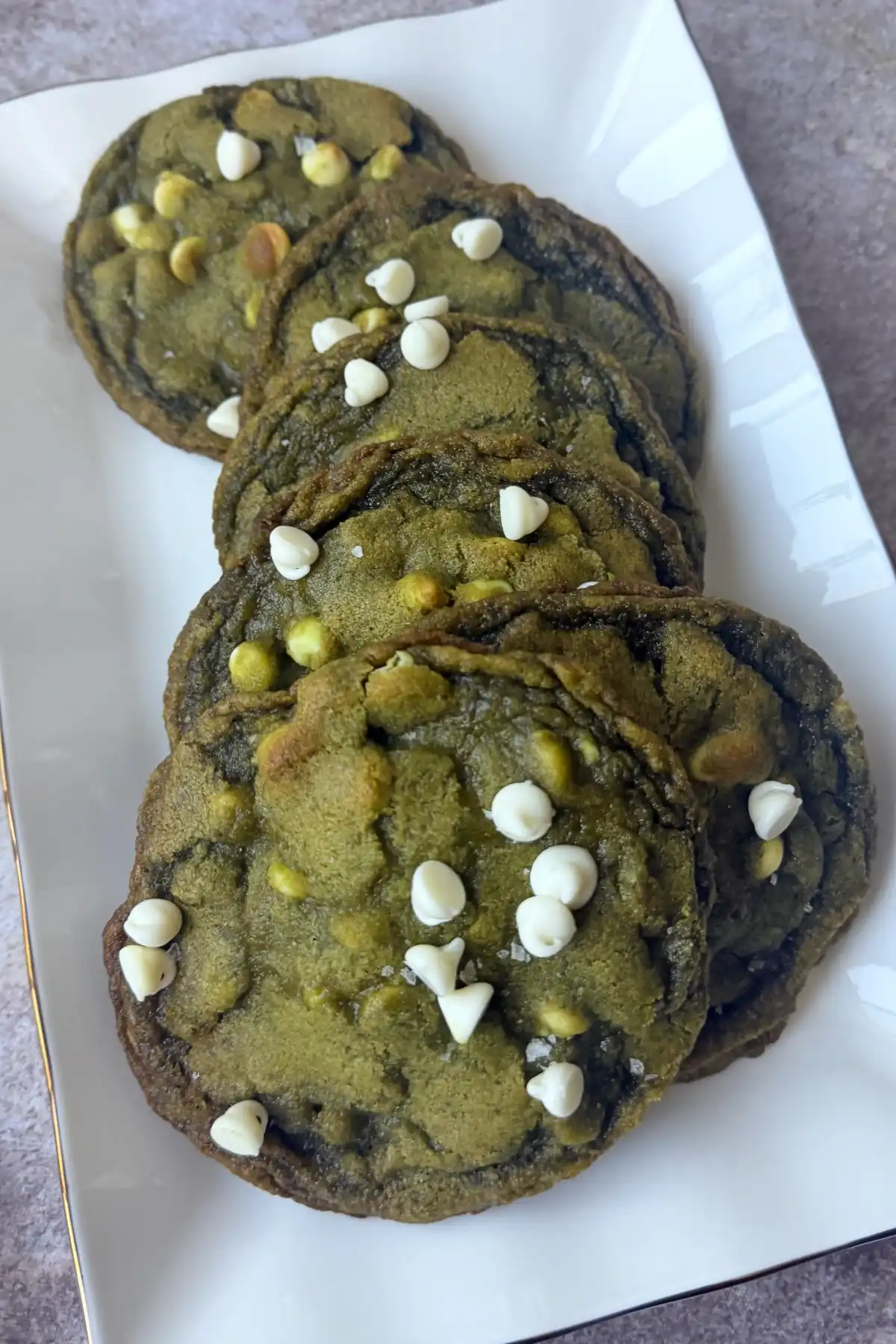 Six matcha white chocolate cookies arranged on a white rectangular plate with extra white chocolate chips on top.