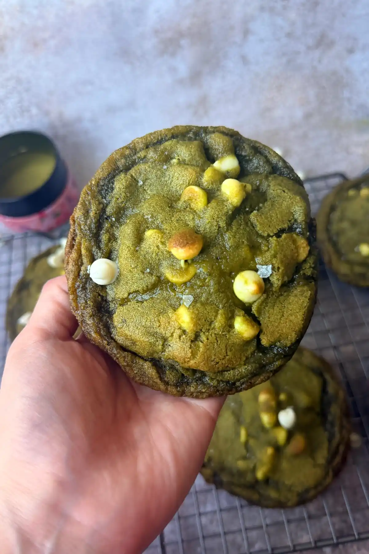 Hand holding a large matcha white chocolate cookie with white chocolate chips and flaky salt, with more cookies in the background.