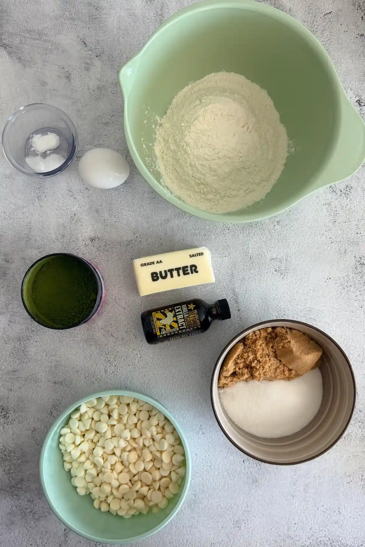 Ingredients for matcha white chocolate cookies arranged on a counter, including flour, butter, egg, vanilla, sugar, matcha powder, salt, baking soda, and white chocolate.
