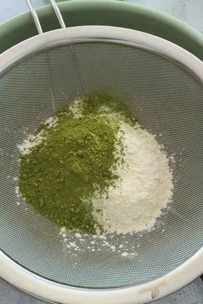 Matcha powder and flour in a fine mesh sieve over a mixing bowl, ready to be sifted.