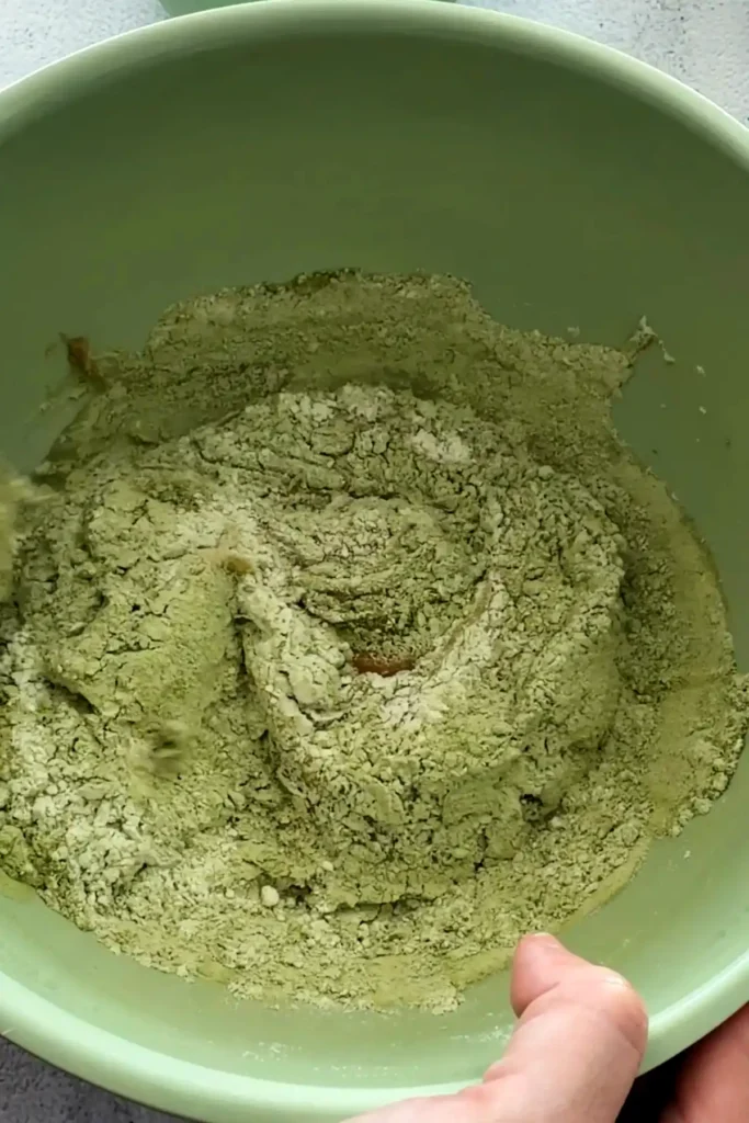 Matcha cookie dough partially mixed in a bowl with dry streaks still visible.