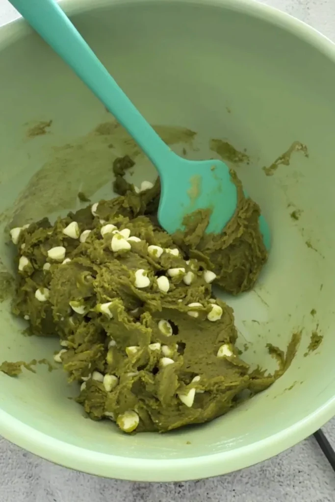 White chocolate chips folded into thick matcha cookie dough in a mixing bowl.