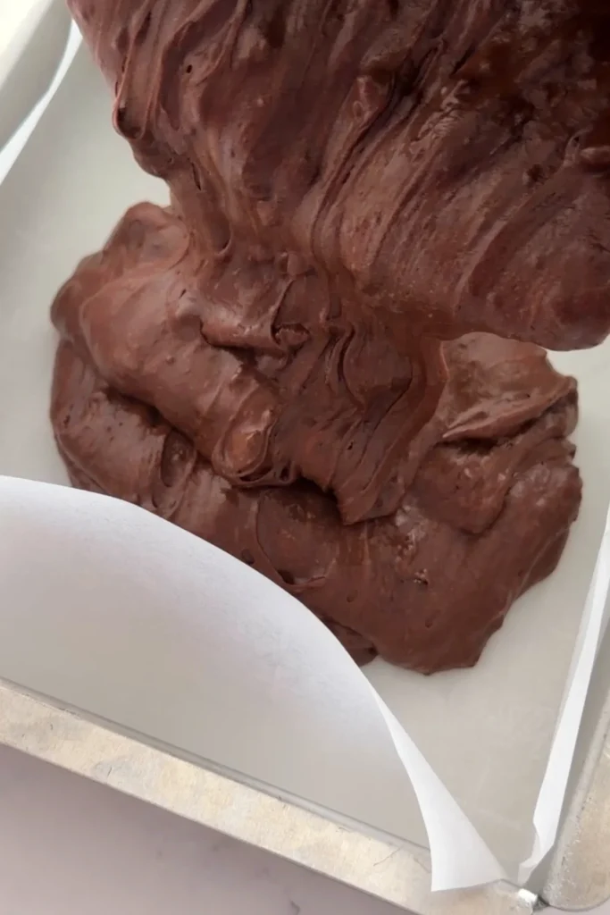 Pouring thick fudgy brownie batter into parchment-lined baking pan