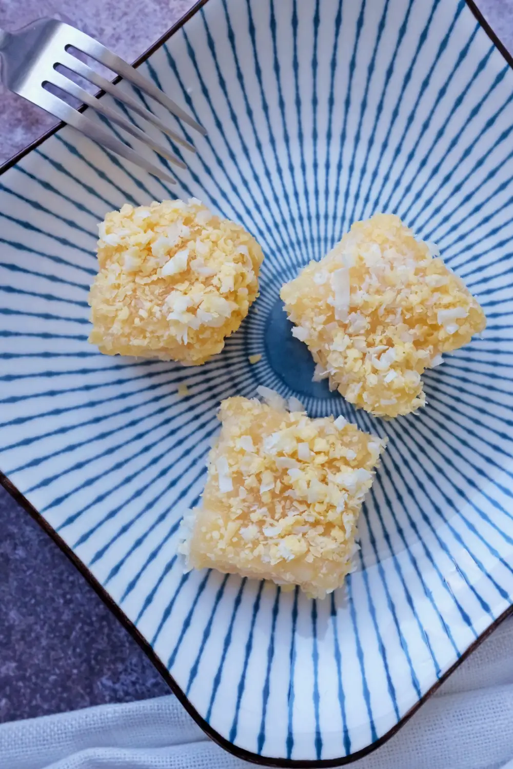 Three pieces of pichi pichi cassava cake topped with grated cheese on a blue patterned plate