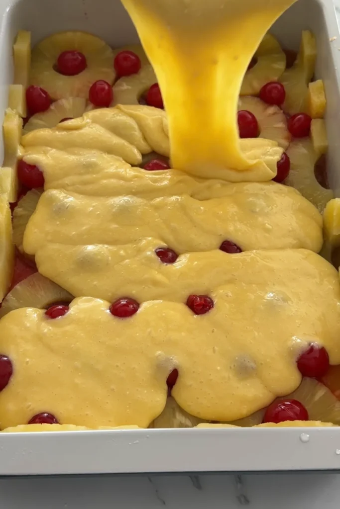Pineapple upside down cake batter being poured over pineapple rings and cherries in a baking dish