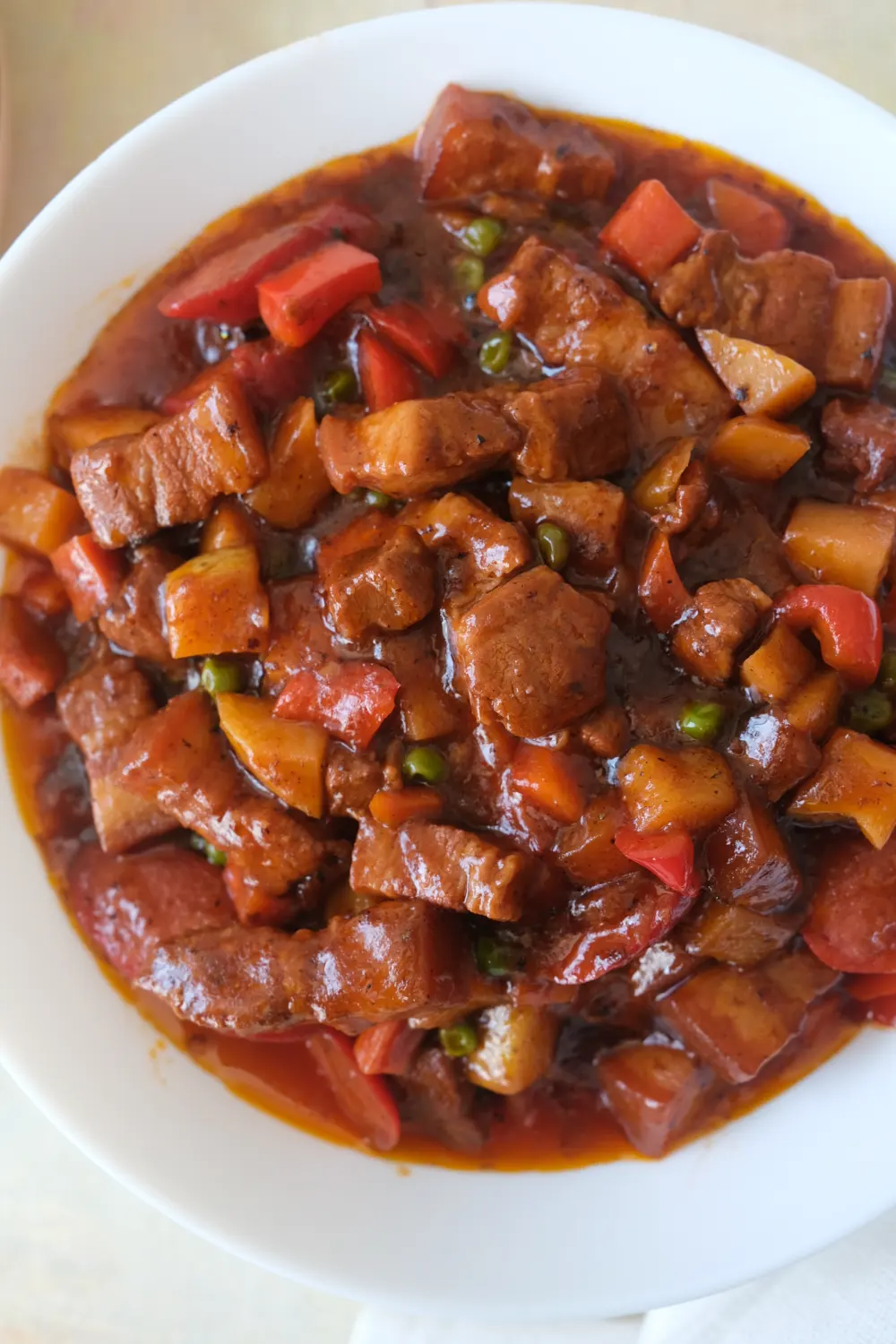 bowl of pork menudo with pork belly in tomato sauce with potatoes, carrots, bell peppers, and peas