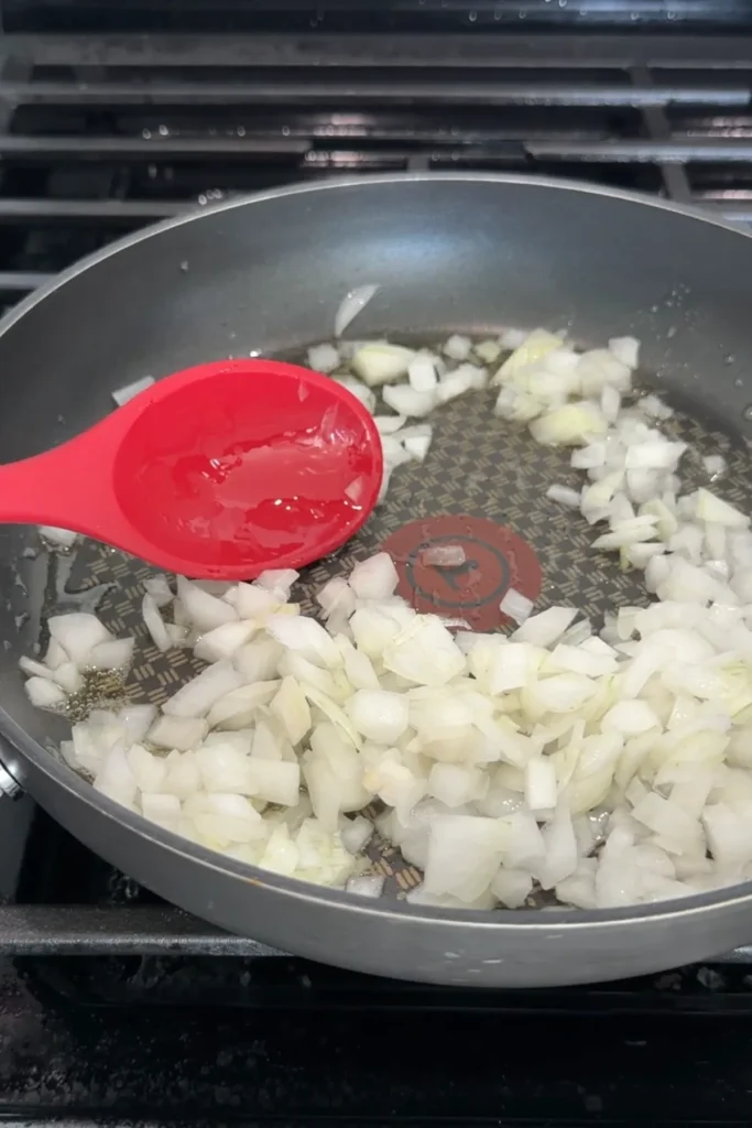 Diced onion cooking in olive oil in a skillet with a red spoon.