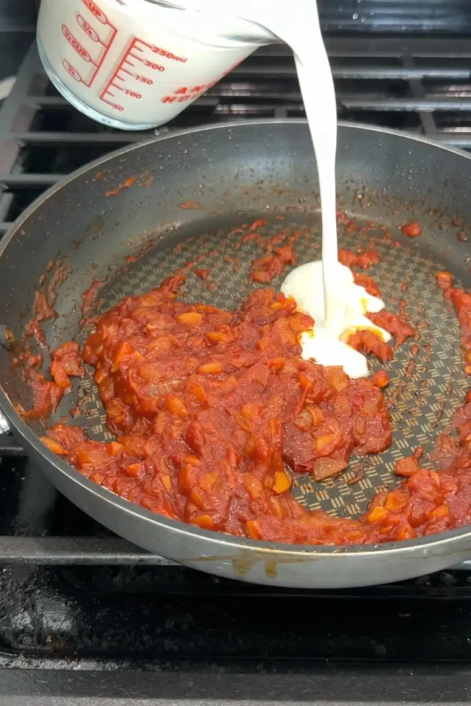 Heavy cream being poured into the cooked tomato paste mixture in a skillet.