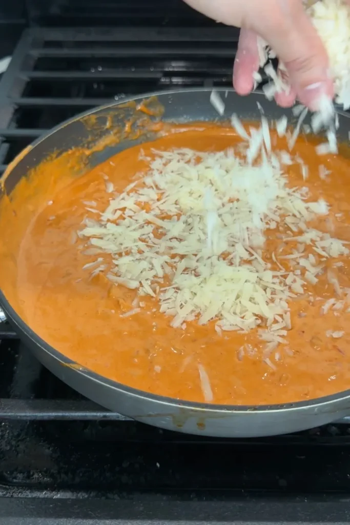 Hand sprinkling grated pecorino into the creamy spicy sauce in a skillet.