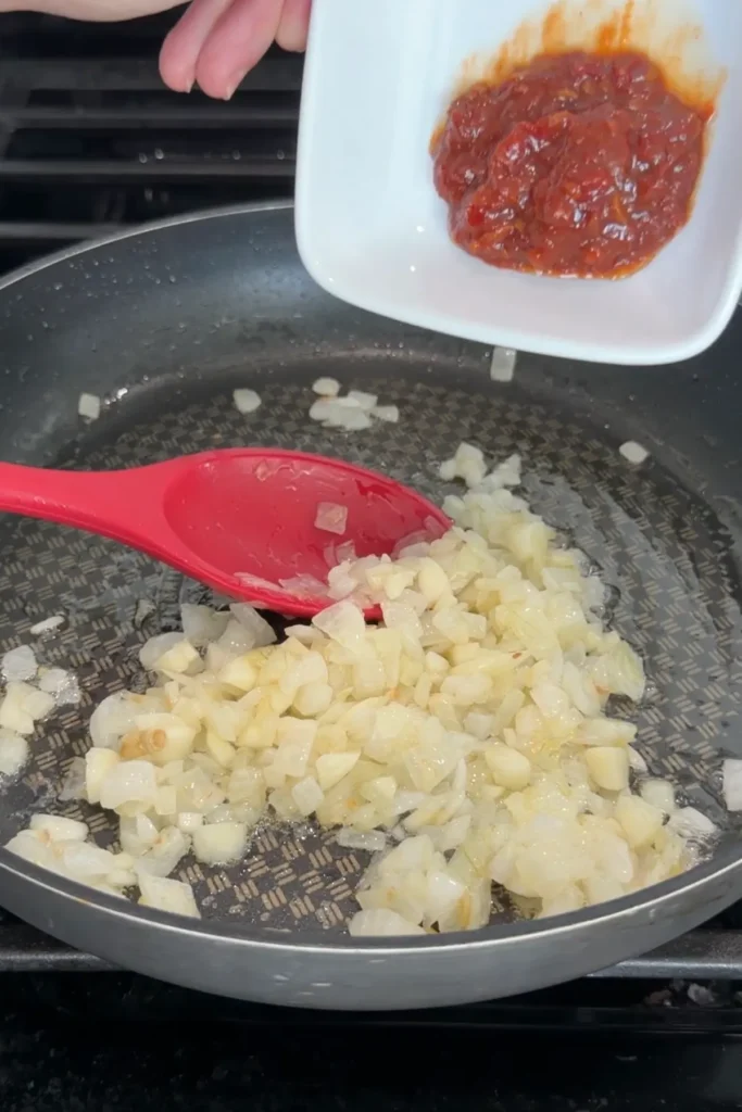 Chili bean sauce being added to softened onion and garlic in a skillet.