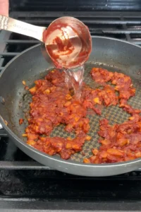 Soju being poured into the cooked tomato paste mixture in a skillet.