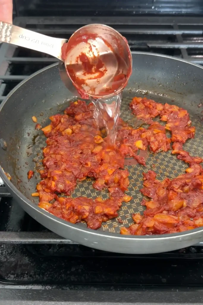 Soju being poured into the cooked tomato paste mixture in a skillet.