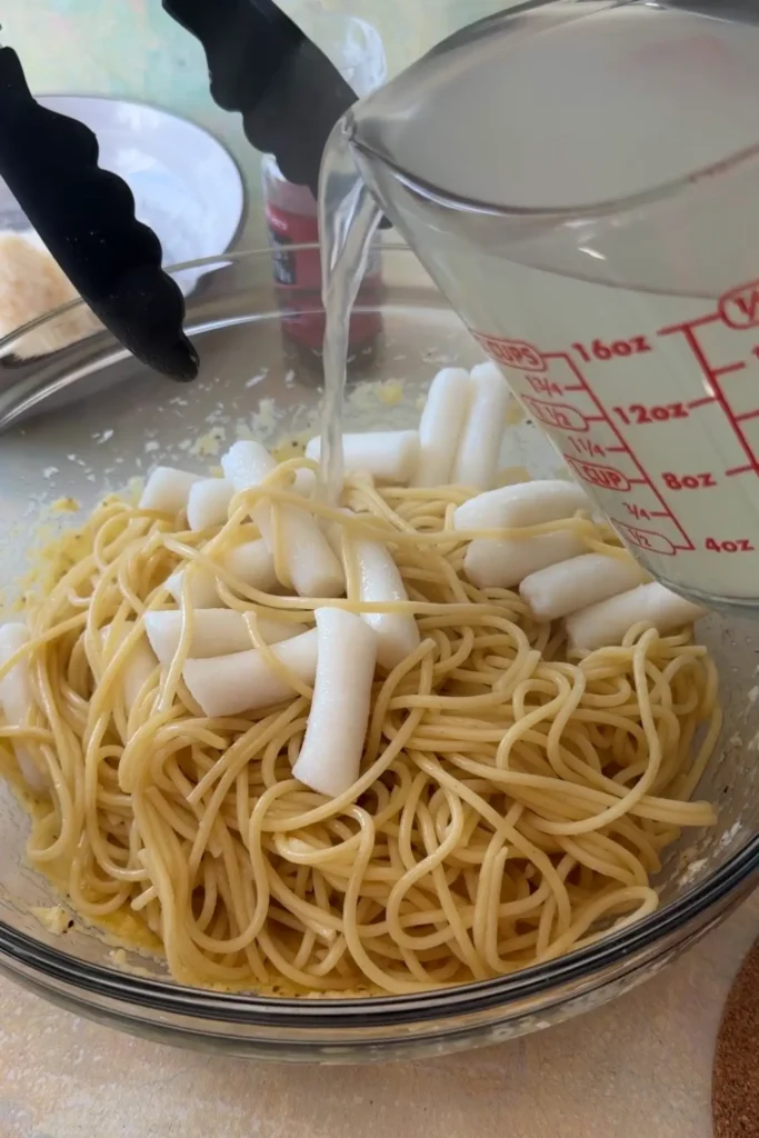 Reserved pasta water being poured over spaghetti and tteokbokki in the carbonara mixture.