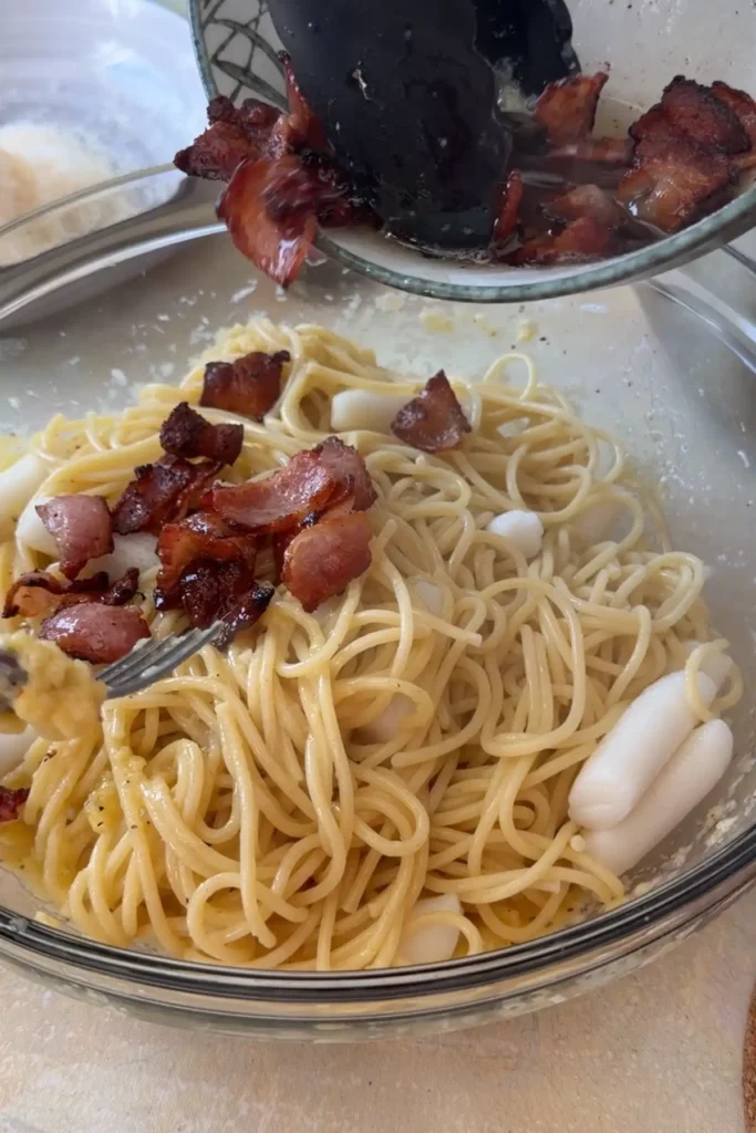 Crispy bacon being added to spaghetti and tteokbokki in the bowl.