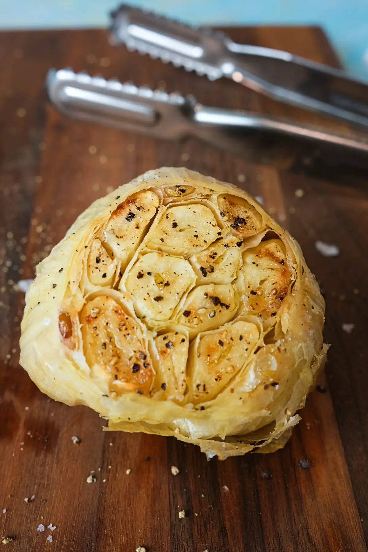 Roasted head of garlic on a wooden cutting board, cut side facing up to show soft golden cloves sprinkled with black pepper and salt, with metal tongs blurred in the background.