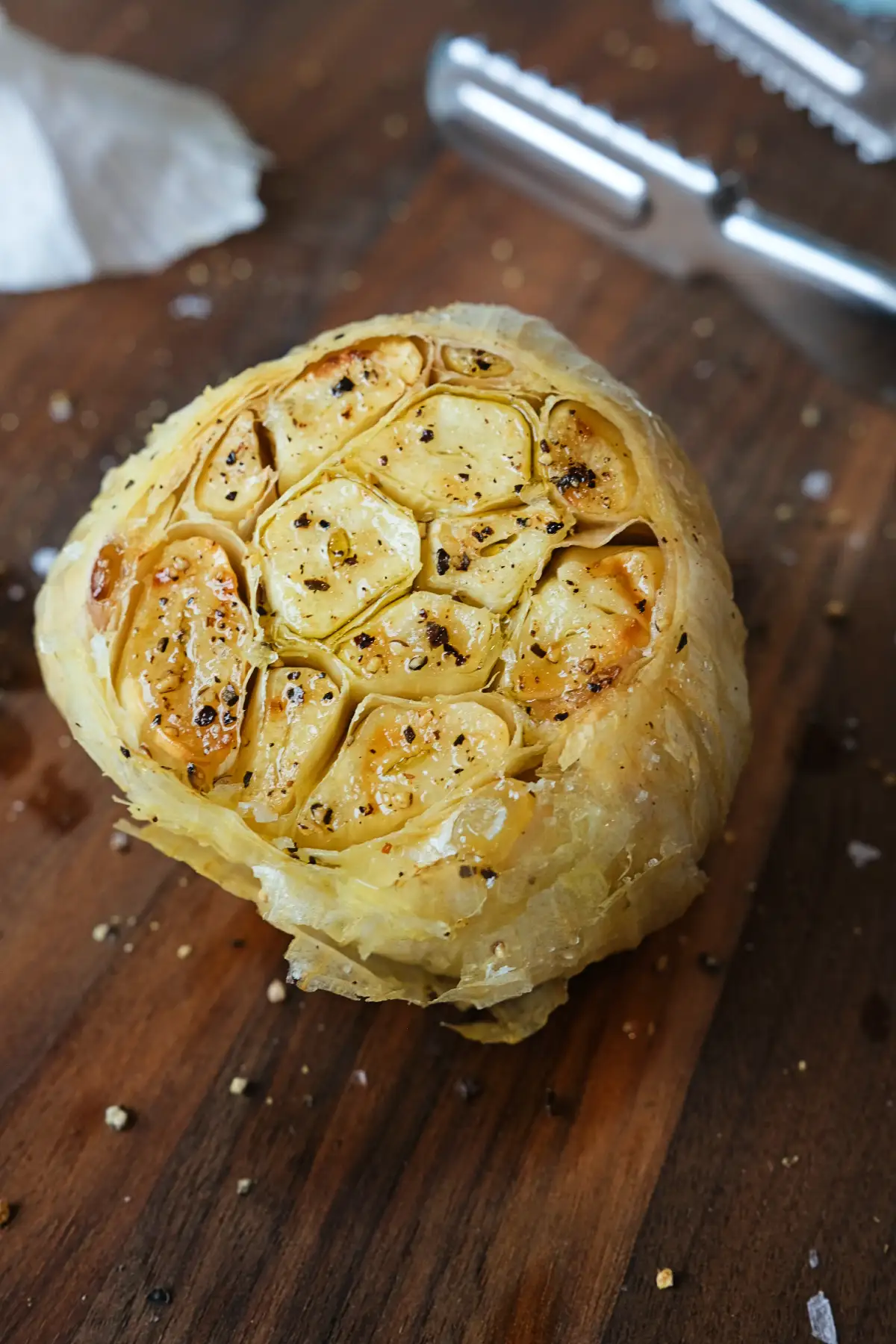 Close view of air fryer roasted garlic resting on a wooden board, with glossy golden cloves exposed and lightly seasoned, while metal tongs sit blurred in the background.