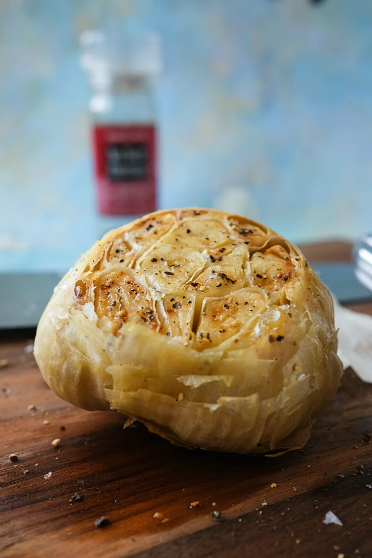 Low-angle shot of a whole roasted garlic head on a wooden cutting board, cut side up and lightly browned, with a seasoning container and kitchen tools softly blurred behind it.