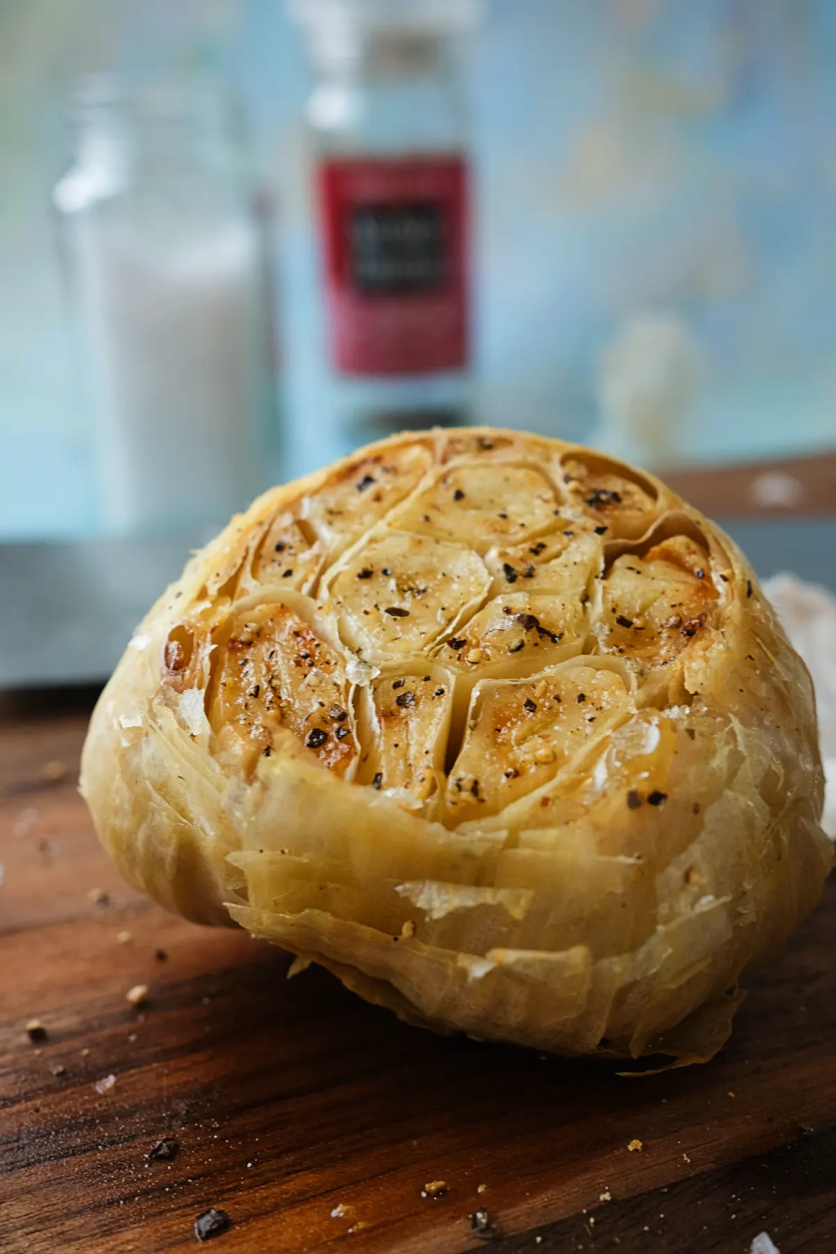 Whole roasted garlic on a wooden cutting board with caramelized, pepper-speckled cloves exposed, framed by blurred salt and seasoning containers in the background.