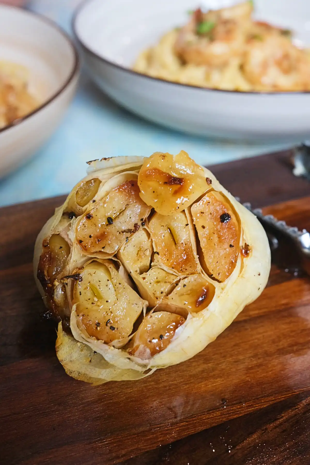Roasted garlic on a wooden board, with shrimp garlic pasta in the background