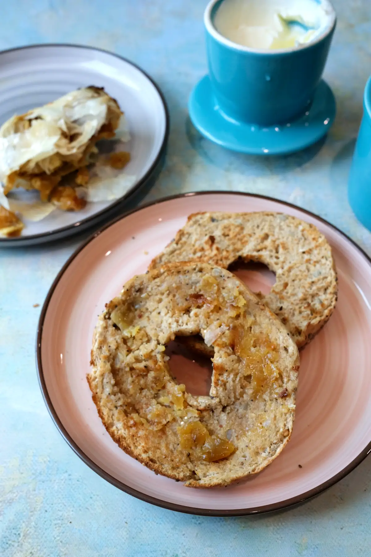 Toasted bagel half on a pink plate spread with soft roasted garlic and melted butter, with the empty top half beside it and a small plate of garlic skins and a butter dish in the background.