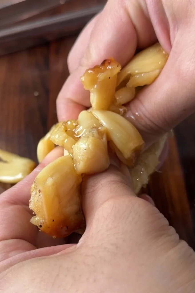 Close-up of hands squeezing roasted garlic cloves out of the softened skin, showing the tender, caramelized cloves slipping out easily.