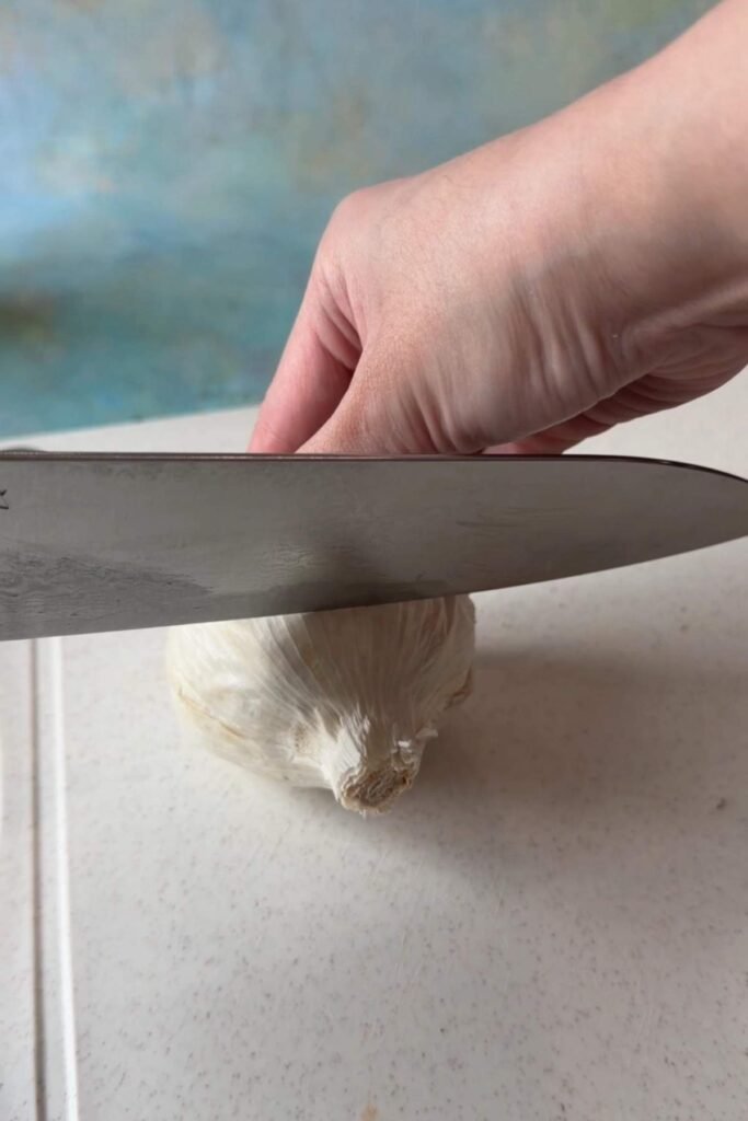 A chef’s knife slicing across the top of a whole raw garlic head on a white cutting board, while one hand holds the garlic steady.