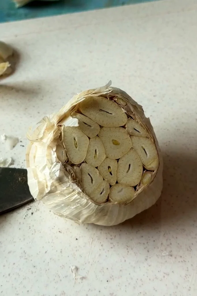 Close-up of a whole garlic head with the top cut off, exposing the raw cloves inside, resting on a white cutting board beside a knife.