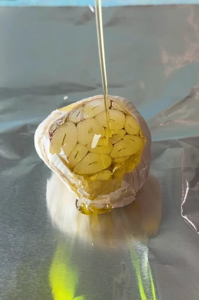 Olive oil being poured over the cut side of a garlic head set on a sheet of foil, coating the exposed cloves with a glossy layer.