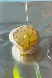 Olive oil being drizzled over a halved head of garlic resting on a sheet of foil, with the cut side facing up and the cloves fully exposed.