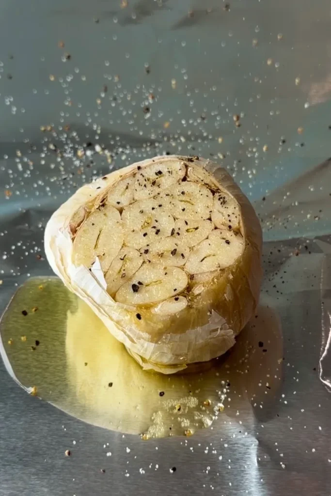 Cut head of garlic on foil sprinkled with salt and black pepper, with oil pooling around the base and seasoning scattered across the cloves.